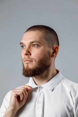 Beyaz tişörtlü yakışıklı bir gencin portresi yakasını dikleştirir ve gri arka planda başka yere bakar. İzole. Kopyalama alanı