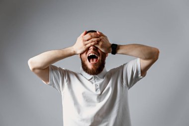 Beyaz tişörtlü mutlu bir gencin portresi. Gri arka planda elleri ile gülüyor ve gözlerini kapıyor. İzole. Kopyalama alanı