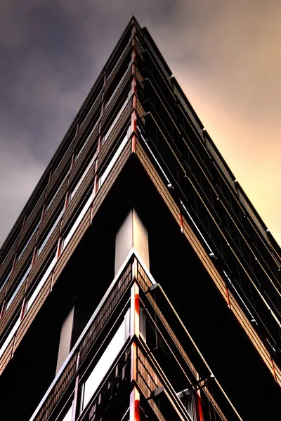 Abstract building pictured in the shape of a triangle. The sky goes from a dark shade on the left to a warmer orange color to the right side of the building. The walls have red shiny metal railings in their sides.