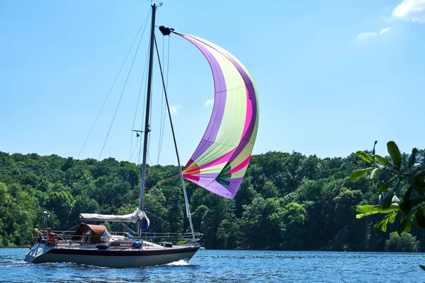 Small yacht with a colorful sail in the front cruising through a lake on a sunny summerday with a forest in the background.