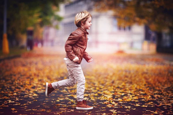 Улыбающийся мальчик, ребенок веселится в осеннем городском парке среди опавших листьев — стоковое фото