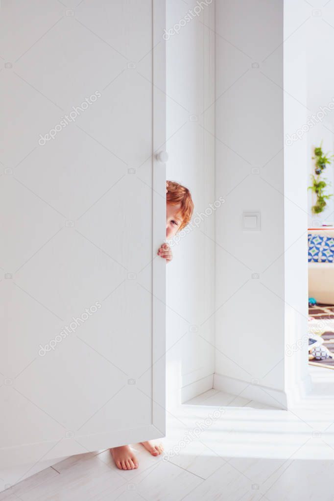 happy toddler baby boy playing hide and seek game, hiding himself behind the cabinet door, candid lifestyle
