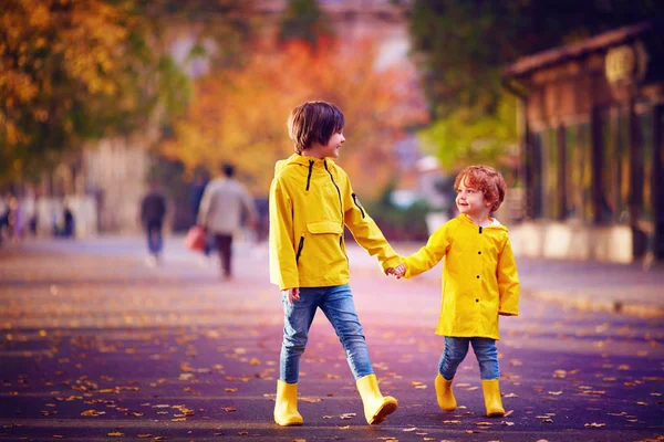 Милые дети, братья держатся за руки, ходят вместе по осенней улице в желтых плащах и сапогах — стоковое фото