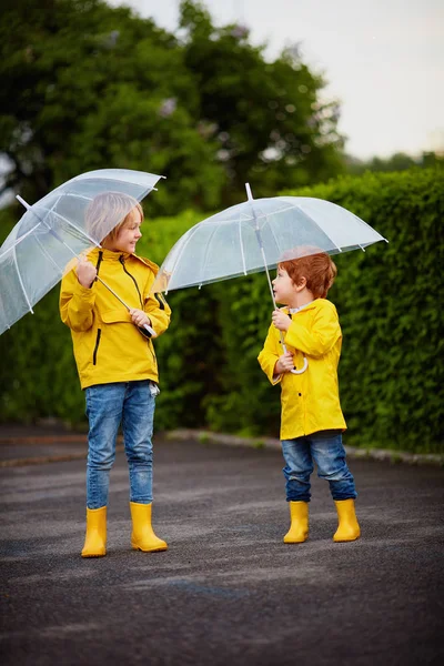 幸せな若い男の子、傘とレインコートとブーツを持つ兄弟は、雨の日に春の公園を歩いて — ストック写真