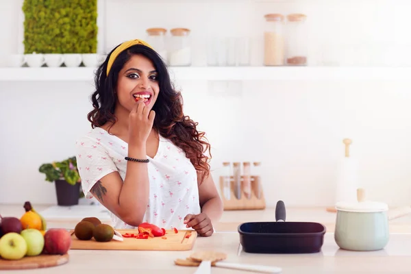 Hermosa mujer joven preparando la comida, mordiendo una verdura durante la cocción — Foto de Stock