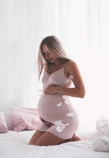 Hermosa mujer embarazada tierna sentada en la cama por la mañana, cogida de la mano en el vientre, concepto de maternidad —  Fotos de Stock