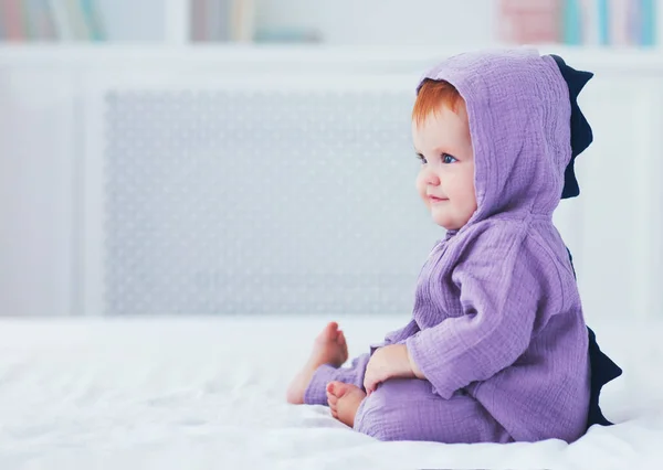 Bela sorrindo, nove meses de idade, bebê bebê menina em traje de dinossauro, está sentado na cama — Fotografia de Stock