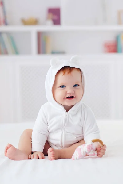 Happy infant baby girl in funny ear hood is sitting on the bed at home — Stock Photo, Image