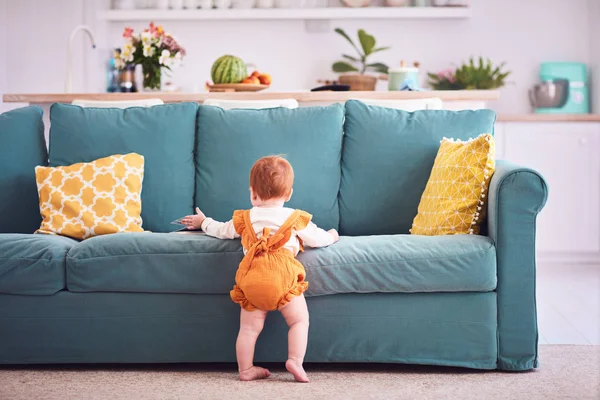 Diez meses de edad, bebé niña en traje de cuerpo amarillo está de pie, apoyado en el sofá en casa —  Fotos de Stock