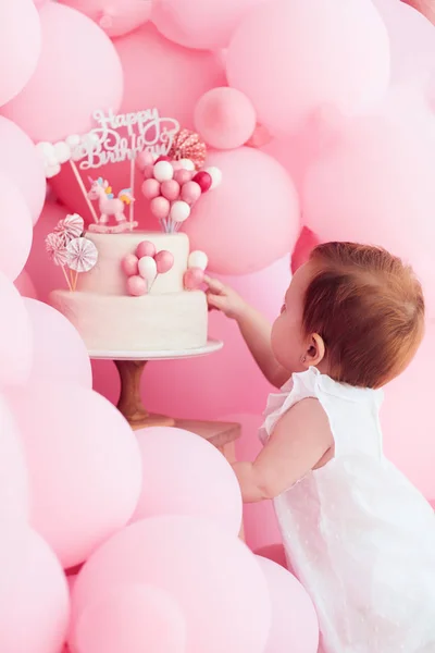 Linda niña metiendo el dedo en el pastel de cumpleaños, decorado con globos de color rosa pastel en la fiesta —  Fotos de Stock