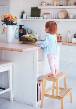 curious infant baby girl trying to reach things on the table in the kitchen with the help of step stool clipart