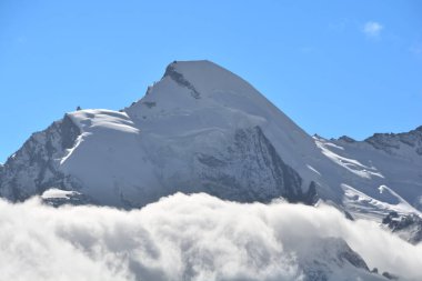 Allalinhorn 'un zirvesi bulutların üzerinde yükseliyor. Sağda İsviçre Alplerindeki Saas Ücreti 'nin üstündeki Mittel Allalin' in dönen buzullu restoranı var.