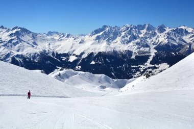 Güneşli bir günde arka planda yüksek bir dağ aralığı olan açık bir kayak pistinde kayak yapan bir insan.