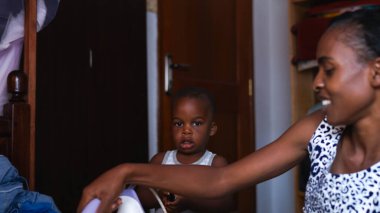 Kampala, Uganda - 01 Temmuz 2017: Aile zamanı, siyah, güzel Afrikalı bir anne, elbiselerini yatak odasına ütülüyor, Afrikalı şirin bir çocuk arkasında akıllı telefon çalıyor, seçici odaklanma, evde kalma 