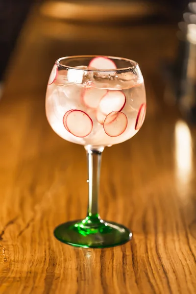 Three colorful cocktails in large wine glasses in a classic bar environment with dozens of blurry liquor bottles in the background.