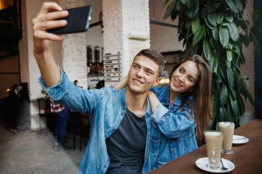 Bir çift kafede kahve içerken fotoğraf çekiyor. Gülümseyen genç adam ve mutlu kadın kahve dükkanında kahve fincanlarıyla otururken cep telefonunda selfie çekiyorlar. Yüksek Çözünürlük.