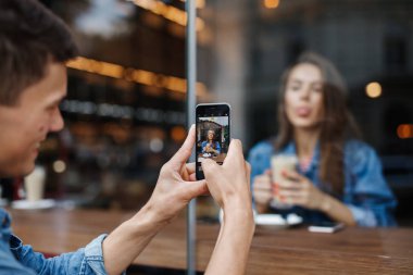 Oğlan telefonda kafedeki pencereden bir kız fotoğrafı çekiyor.