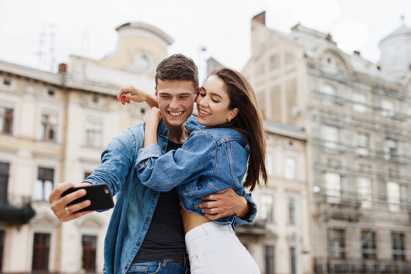 Couple Taking Photos In  big city street. Smiling Young Man And Happy Woman Doing Selfie Photo On Mobile Phone. High Resolution.