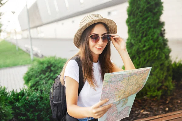 Young smiling  traveler girl with coffee searching location on map sitting on bench. during a summer trave