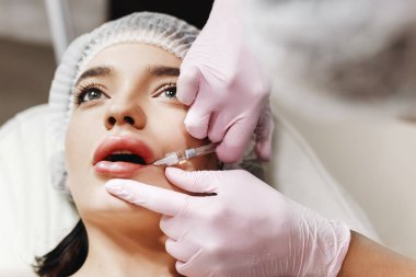 Dudak geliştirme için hazırlık süreci. Doktor hastanın önünde hyaluronik asit yukarı çekiyor. Çok güzel bir yüzün özel şapka ve bir doktor pembe eldiven eller genç kızla