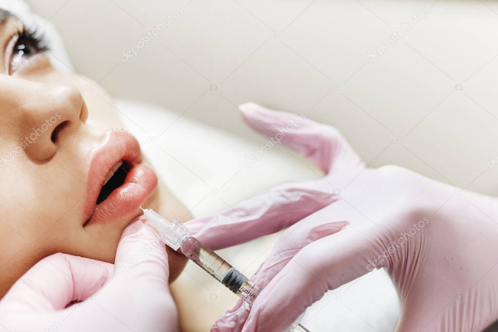 The process of lips enhancement. Cosmetician makes an injection of hyaluronic acid in a beautiful lips. The young girl with a beautiful face in the special hat and hands of the doctor in the pink gloves.