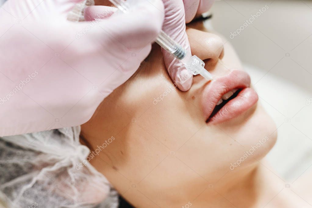 The process of lips enhancement. Cosmetician makes an injection of hyaluronic acid in a beautiful lips. The young girl with a beautiful face in the special hat and hands of the doctor in the pink gloves.