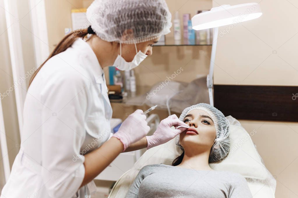 The process of lips enhancement. The doctor reviews enhanced lips. The young girl with a beautiful face in special hat and  the hands of the doctor in pink gloves.