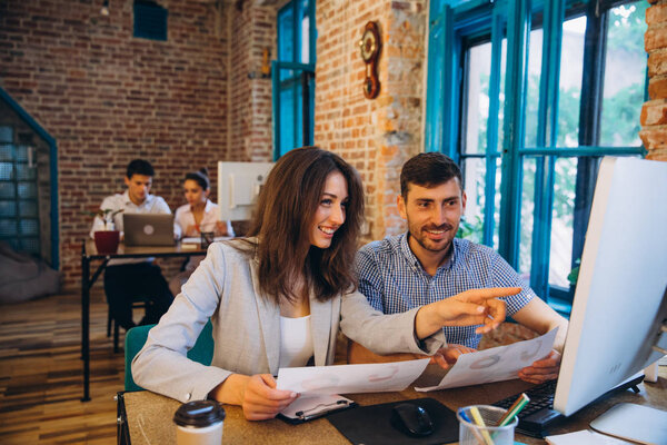 Двое молодых сотрудников работают за ноутбуком в солнечном офисе. Женщина держит бумажные документы и указывает на экран ноутбука. Размытый бэкграунд.
