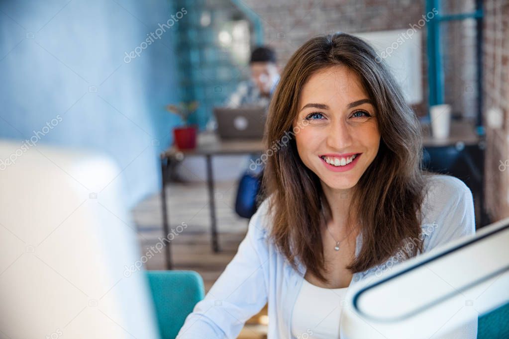 Attractive businesswoman working on laptop and smiling for camera in workplace, successful confident executive manager sitting at office desk, young e-business owner posing at work, headshot portrait.