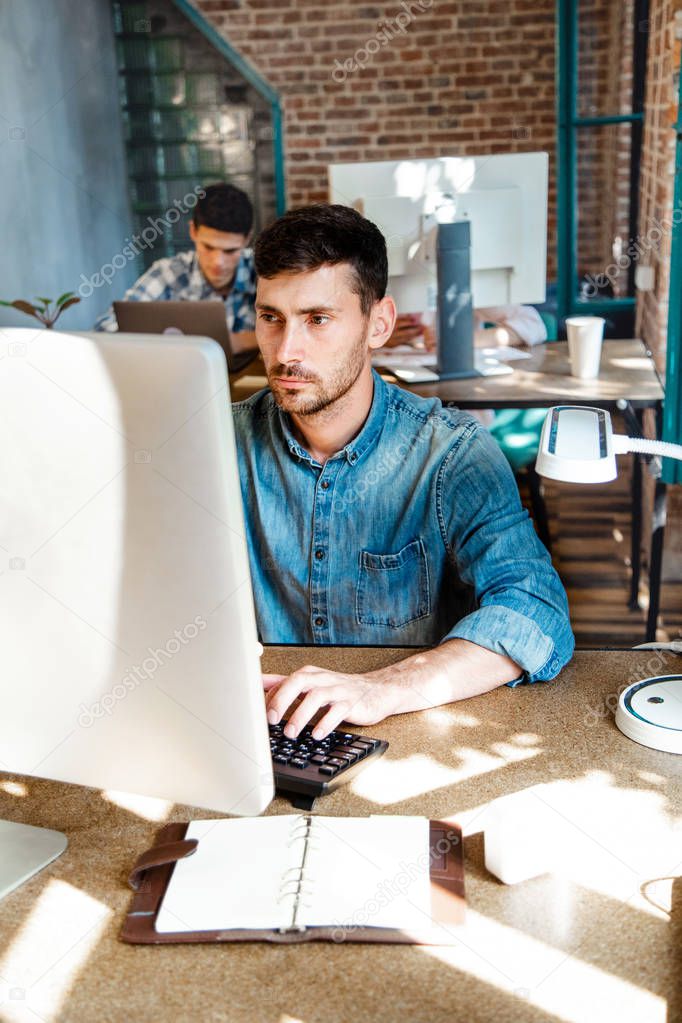 Young bearded businessman in denim shirt is sitting in office at table and is using laptop with charts, graphs and diagrams on screen. Man working.startu