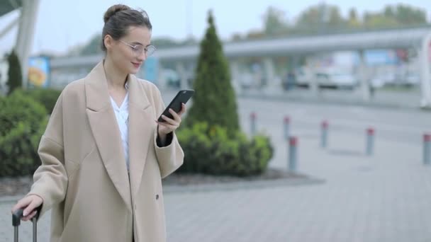 空港ターミナルで待っているゲートでスマート フォンで女性 スマート フォン 機内持ち込み手荷物のトロリーの話に座っている若いカジュアルなビジネス女性の空気旅行の概念 若く美しい — ストック動画