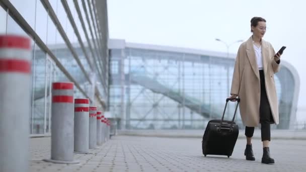 机场妇女在智能电话在门等待在终端 航空旅行理念与年轻的休闲商务女性坐在一起谈论智能手机 随身携带手提行李推车 美丽的年轻 — 图库视频影像