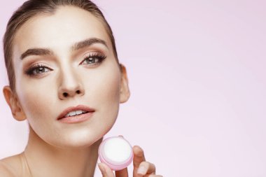 Mükemmel çıplak makyajlı kız güzellik portresi. Krem basılı tutup yüzünü dokunaklı. Pembe bir arka plan. Güzellik Salonu. Baş ve omuzlar, stüdyo, kapalı 