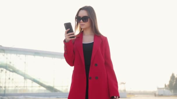 空港ターミナルで待っているゲートでスマート フォンで女性 スマート フォン 機内持ち込み手荷物のトロリーの話に座っている若いカジュアルなビジネス女性の空気旅行の概念 若く美しい — ストック動画