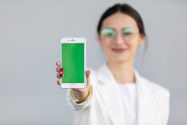 Beyaz ceketli gülümseyen genç iş kadını yeşil ekranlı akıllı telefonu gösteriyor, uygulama