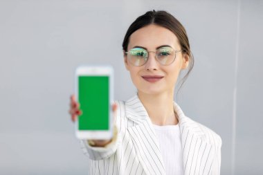 Beyaz ceketli gülümseyen genç iş kadını yeşil ekranlı akıllı telefonu gösteriyor, uygulama