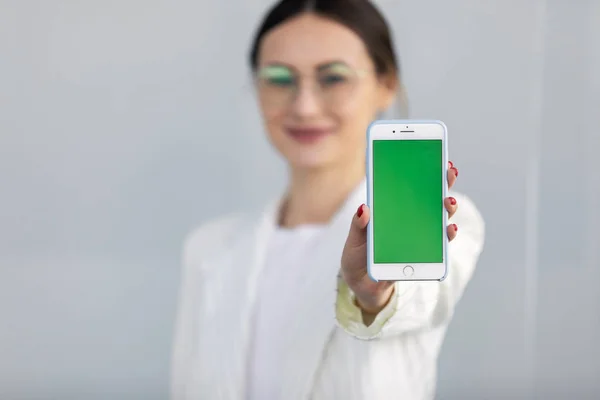 Beyaz ceketli gülümseyen genç iş kadını yeşil ekranlı akıllı telefonu gösteriyor, uygulama