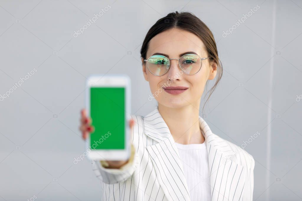 Smiling young business woman in white jacket  showing smartphone with green screen, app