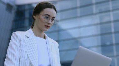 Modern iş merkezinde çalışırken dizüstü bilgisayarını dışarıdan kullanan genç profesyonel iş kadınının yakın çekimi, arka planda modern binalar, parlama ışığı, film efektleri, yavaş çekim