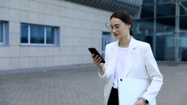 Beyaz gömlek giymiş ve şehrin dışında yavaş hareket cep telefonu kısa mesaj yazarak profesyonel kadın işveren kopma yürürken modern akıllı telefon kullanarak güzel genç iş kadını
