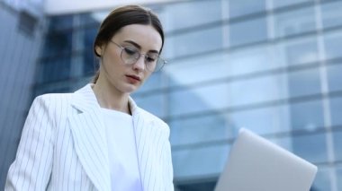 Modern iş merkezinde çalışırken dizüstü bilgisayarını dışarıdan kullanan genç profesyonel iş kadınının yakın çekimi, arka planda modern binalar, parlama ışığı, film efektleri, yavaş çekim