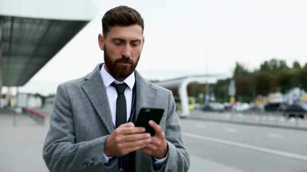 スマートフォンを使って空港で笑顔を見せているビジネスマン 空港で携帯電話を持つ若いビジネスエグゼクティブ — ストック動画