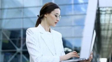 Modern iş merkezinde çalışırken dizüstü bilgisayarını dışarıdan kullanan genç profesyonel iş kadınının yakın çekimi, arka planda modern binalar, parlama ışığı, film efektleri, yavaş çekim