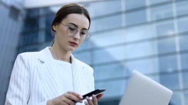 Modern iş merkezinde çalışırken dizüstü bilgisayarını dışarıdan kullanan genç profesyonel iş kadınının yakın çekimi, arka planda modern binalar, parlama ışığı, film efektleri, yavaş çekim