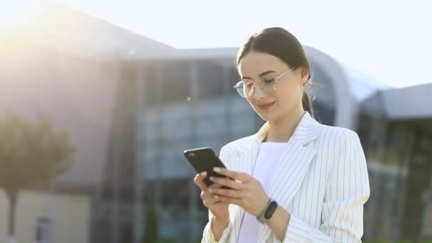 美丽的年轻女商人穿着白衬衫和步行在城市 在外面 慢动作手机上键入文本消息的专业女雇主伸长时使用现代的智能手机 — 图库视频影像