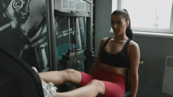 Hermosa mujer joven en sujetador deportivo y polainas respirando profundamente mientras hace ejercicios para las piernas en la máquina de fitness. Chica fuerte con cabello oscuro usando pesas durante el entrenamiento en el gimnasio . — Vídeos de Stock