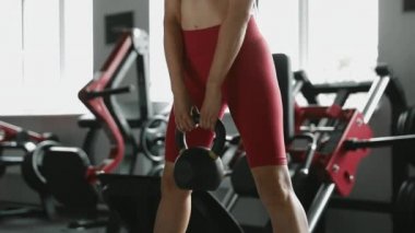 Siyah saçlı çekici kaslı kadın modern spor salonunda siyah halterle egzersiz yapıyor. Spor kıyafetleri giyen genç bir kız. Bacakları içeride ağırlık çalışıyor..