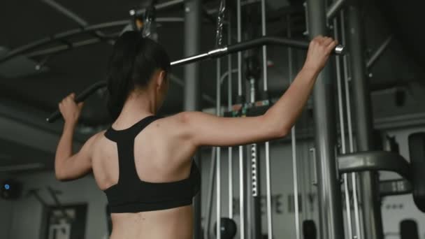 現代のジムでプルダウンマシン上で運動暗い髪を持つ強い若い女性のバックビュー アクティブガールトレーニング上背部と腕でスポーツ機器 — ストック動画