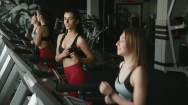 Spor kıyafetleriyle spor salonunda koşu bandında koşan iki mutlu aktif kız. Güzel bayan arkadaşlar sohbet ediyor ve birlikte kardiyo yapıyorlar. Dayanıklılık ve motivasyon kavramı.