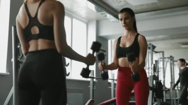 Spor kıyafetleri içinde iki güzel kadın spor salonunda ağır dambıllarla antrenman yapıyor. Mutlu bayan arkadaşlar kaslarını içeride şişiriyorlar..